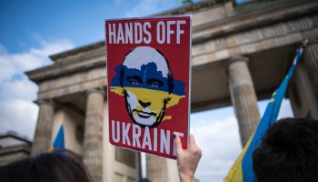 A protester holds up a poster reading "Hands Off Ukraine" and depicting the face of Russian President Vladimir Putin covered with a map of Ukraine in the colors of the Ukrainian flag during a peace demonstration in front of the Brandenburger Gate in Berlin on February 19, 2022.