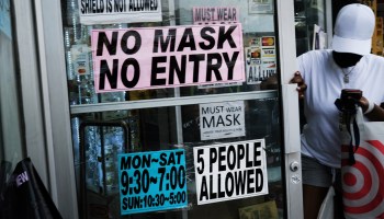 A person opens a door to a shop. The door has signs that describe the hours and say, "No mask, no entry" and "5 people allowed."