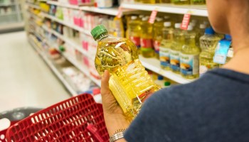 A woman holds a bottle of cooking oil in a grocery store aisle.