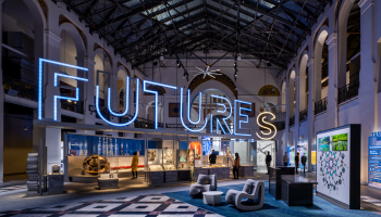The entrance hall for FUTURES at the Smithsonian Arts and Industries Building. It includes a neon sign that says, "Futures," as well as some chairs, and people walking through the large main hall of the building.