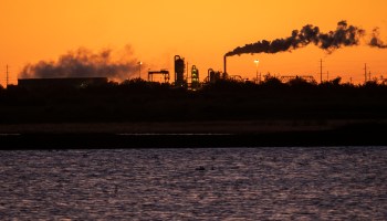 Smoke stacks emit pollution behind a body of water during sunset.