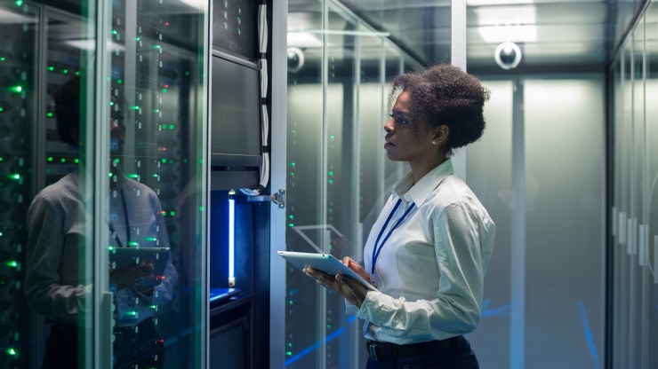 A woman working on a tablet examines a rack of servers in a data center.