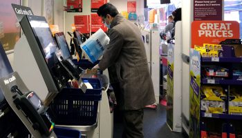 A shopper wearing a face mask loads some toilet paper into a bag at a self-checkout kiosk in a supermarket.