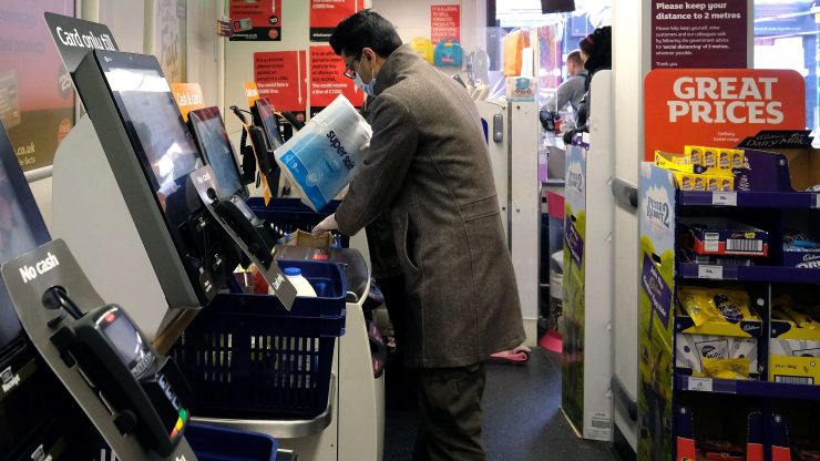 A shopper wearing a face mask loads some toilet paper into a bag at a self-checkout kiosk in a supermarket.