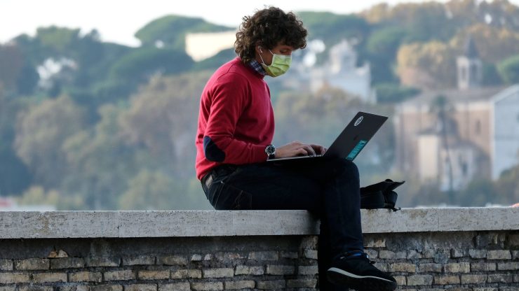 A man wearing a surgical face mask and red long-sleeved shirt sits on a walled fence with his back facing a landscape of trees and buildings, as he uses his laptop.