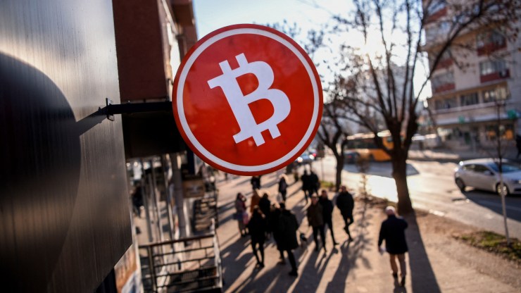 Pedestrians walk under a Bitcoin cryptocurrency symbol displayed at a cryptocurrency exchange shop in Pristina on January 17, 2022. - Kosovo police on January 8 seized hundreds of cryptocurrency mining machines and arrested one person in the tense ethnic-Serb majority north as the country suffered an energy crisis. Cryptocurrencies like Bitcoin are created through solving complex equations -- an endeavour that consumes enormous amounts of energy. Tensions between the Serb-majority area and the ethnic Albanian majority government are running high and Kosovo's government on Tuesday brought in a temporary ban on cryptocurrency mining in an effort to bring down electricity consumption. (Photo by Armend NIMANI / AFP) (Photo by ARMEND NIMANI/AFP via Getty Images)