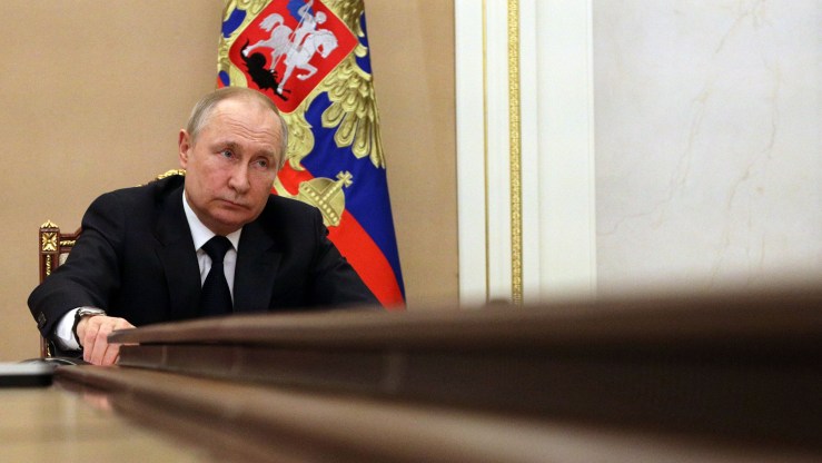 Vladimir Putin sits at a table with a flag behind him.