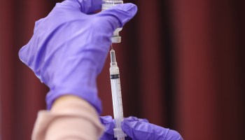 A hand in a latex medical glove fills a syringe from a vaccine vial.