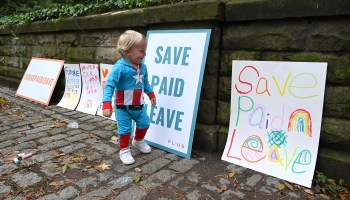 A child in a superhero costume runs past signs that say "Save Paid Leave."