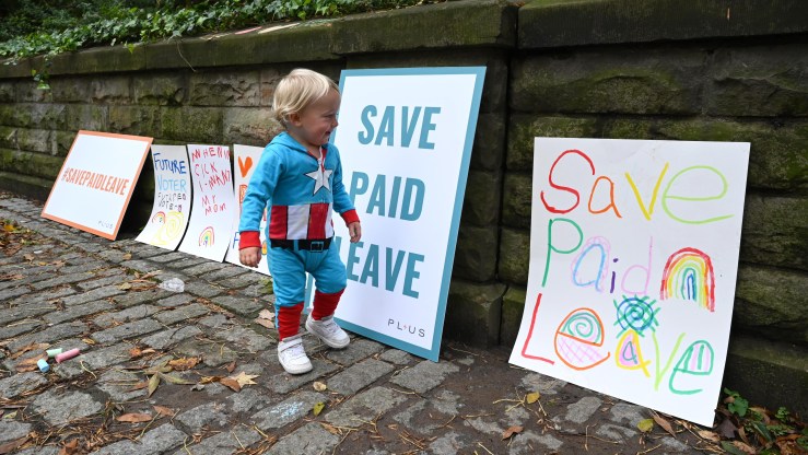A child in a superhero costume runs past signs that say "Save Paid Leave."