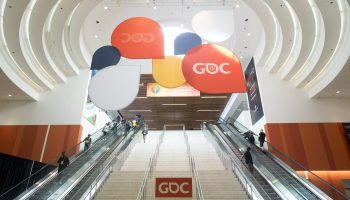 A set of escalators lead up to a second floor. The ceiling is white and curved, and includes a sign post with multicolor red, yellow, and blue circles that read: "GDC."