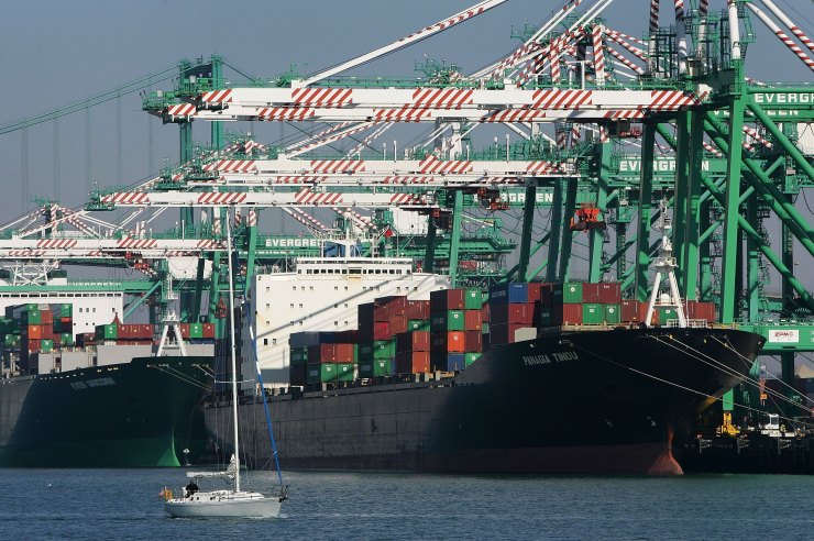 A cargo ship loaded with shipping containers. A much smaller sailing boat is in the foreground.