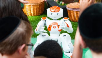 A short orange and white robot with arms and legs looks at the camera. In the foreground, as the backs of heads of kids interacting with it.