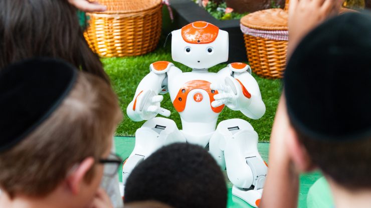 A short orange and white robot with arms and legs looks at the camera. In the foreground, as the backs of heads of kids interacting with it.