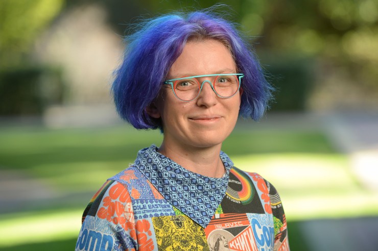 Quinn Dombrowski smiles with blue/purple hair and a colorful shirt in an outdoor setting.