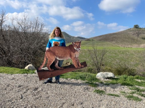 Beth Pratt is pictured here on the north side of Highway 101 with her cardboard cutout of P22 – the mountain lion who is the inspiration behind the $90 million wildlife crossing being built in Agoura Hills. Behind her is where the wildlife crossing will be located.