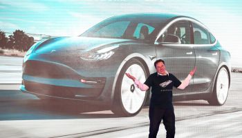 Tesla founder Elon Musk stands with his arms in the air in front of a large picture of a black Tesla.