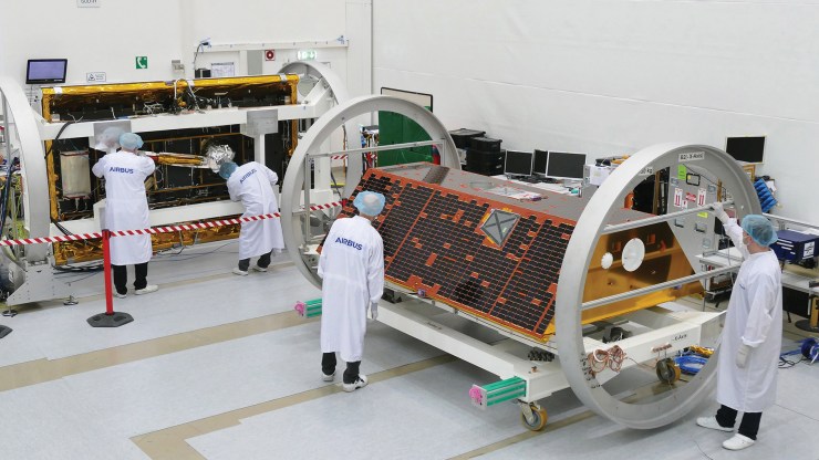 Four scientists wearing lab coats, masks and hair nets, work on the trapezoid-shaped GRACE-Follow On satellites in a laboratory.