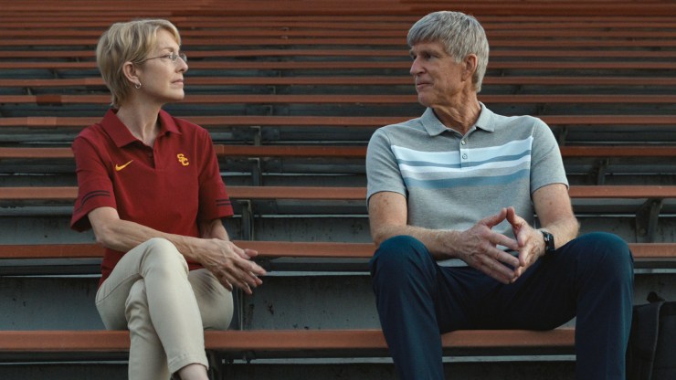 A woman wearing a University of Southern California polo shirt sits in the bleachers near a sports field. She's sitting next to an actor portraying Rick Singer in a production still from the documentary "Operation Varsity Blues: The College Admissions Scandal."
