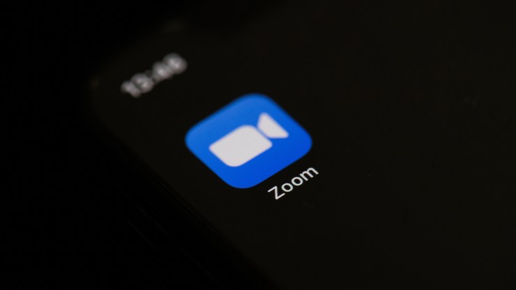 The logo for Zoom is seen on a cell phone in front of a dark background.