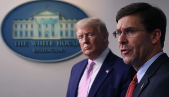 Mark Esper and Donald Trump stand shoulder to shoulder, slightly off to the side. On the wall is the White House insignia.