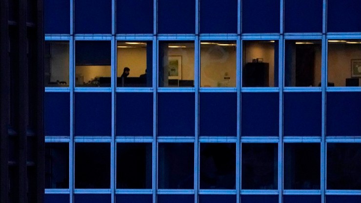 The silhouette of an employee is seen in a row of windows in a high-rise office building. All other floors are dark and empty.