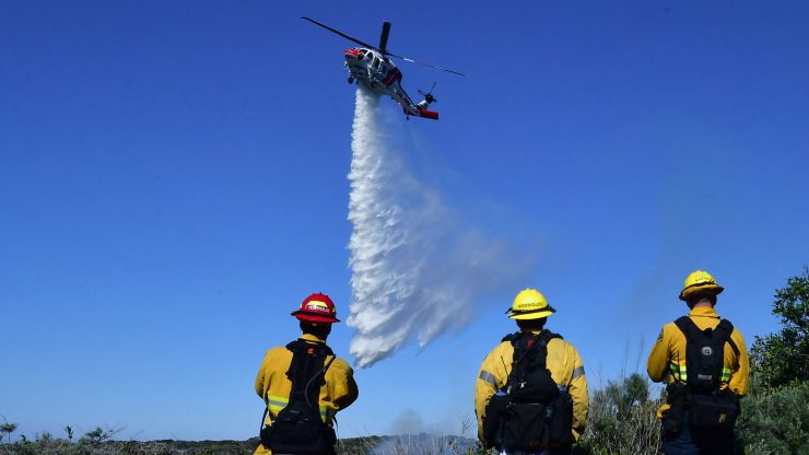 Three firefighters watching as a helicopter releases water onto a fire during the day.