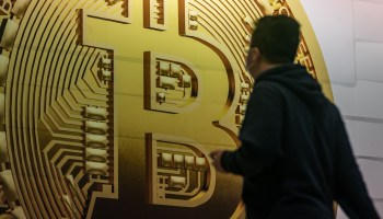 A person stands in front of a large gold bitcoin token.