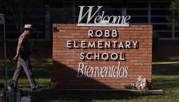 An officer walks past a sign that reads "Robb Elementary School, Welcome, Bienvenidos." A bouquet of flower rests at the sign's base.