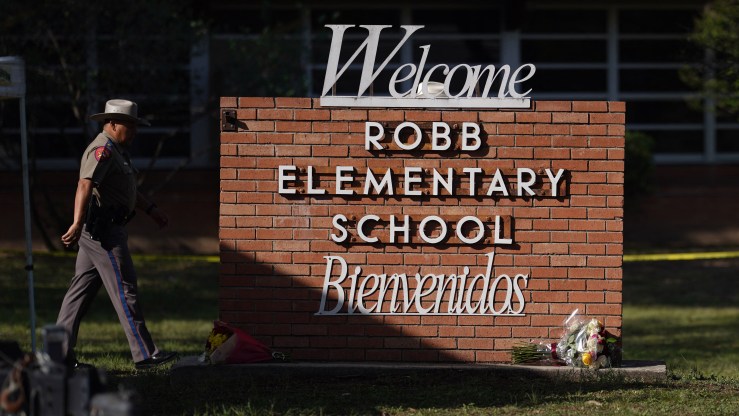 An officer walks past a sign that reads "Robb Elementary School, Welcome, Bienvenidos." A bouquet of flower rests at the sign's base.