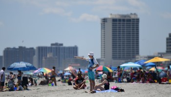 A man in a swimsuit and bucket hat points toward the waterfront in front of dozens of other beachgoers.