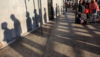 The long shadows of people waiting in line to cross the U.S. border are stretched across the sidewalk and the wall of a building.