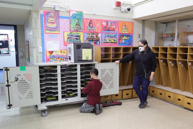 A female teacher stands at the side of a laptop cart in a classroom while one of her students pulls a computer off the shelves.
