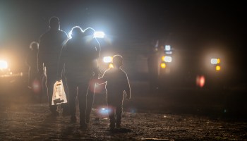 A family of migrants walks toward border patrol cars. Their figures are hazily illuminated by the vehicle headlights in the night.
