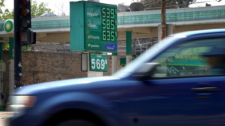 A sign displays gas prices at a gas station. A blue car drives by in the foreground.