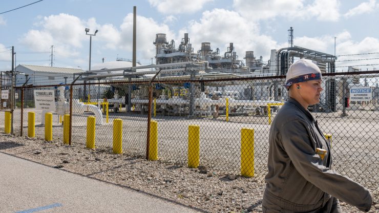 The profile of a person walking in front of an oil refinery in Texas on a sunny day.