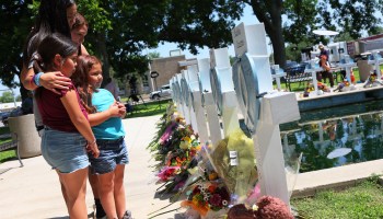 Four young girls visit a memorial, listing all of the people killed at the Uvalde shooting. They look at crosses with bouquets of flowers resting at the base of each.