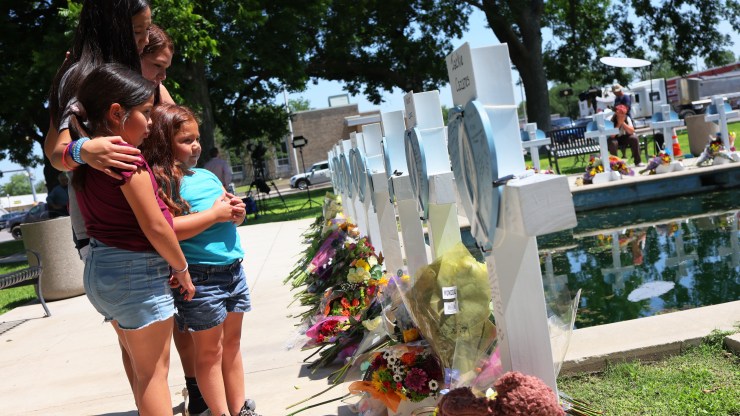 Four young girls visit a memorial, listing all of the people killed at the Uvalde shooting. They look at crosses with bouquets of flowers resting at the base of each.