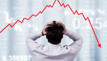 A man clutches his head in stress looking at stock market prices falling, shown by a dramatic red arrow pointing down.