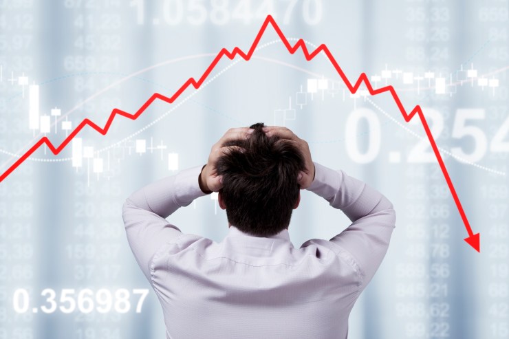 A man clutches his head in stress looking at stock market prices falling, shown by a dramatic red arrow pointing down.