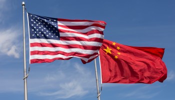 An American and Chinese flag are seen side-by-side on flagpoles during a sunny day.