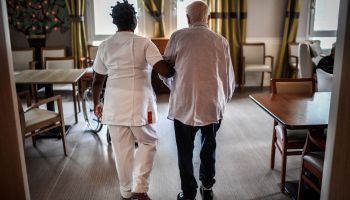 An elderly resident walks in a residential facility with the help of a nurse.