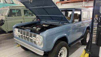 A light blue Ford Bronco is in a car shop with its hood popped open.