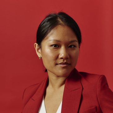 A headshot of journalist Karen Hao wearing a red coat with a red backdrop.