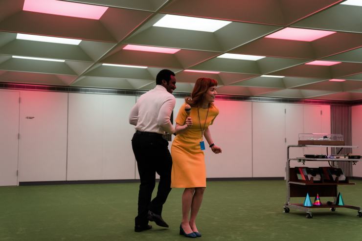 Two office workers dance inside an office room dimly lit by red and white lights. Party hats, music discs and other decorations sit on a rolling cart nearby.