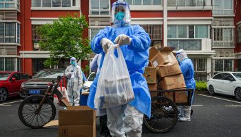 A volunteer in a protective mask, gown and gloves holds a bag of food near a motorcycle that carries other foods and goods.