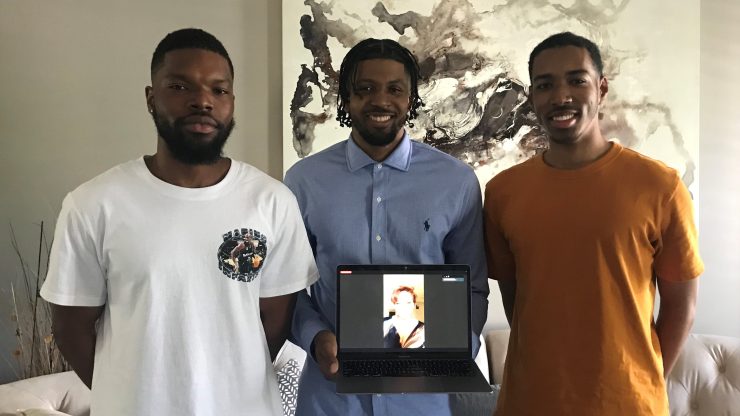 Tobi Plumpter, Guy DeWeever II and Emanuel Perez smile in a line and hold a laptop that shows Leah Smith. They are in a living room in front of a piece of art.