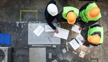 Engineer workers wearing hard hats and neon vests are seen from above examining a factory table of documents.