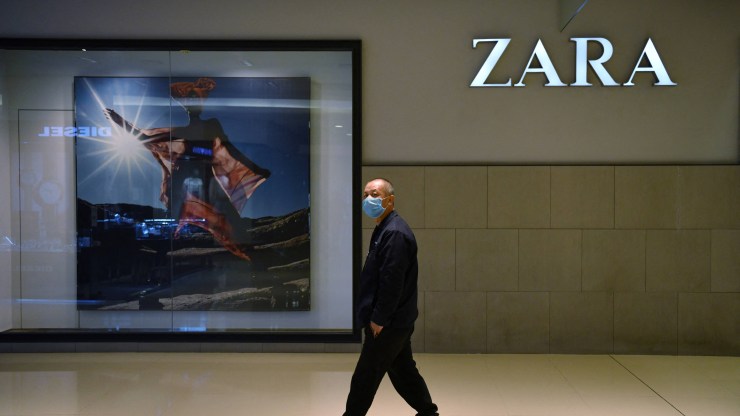 A masked man walks past a Zara store. On the left is an advertisement of a woman wearing Zara clothing and on the right is the name of the store.