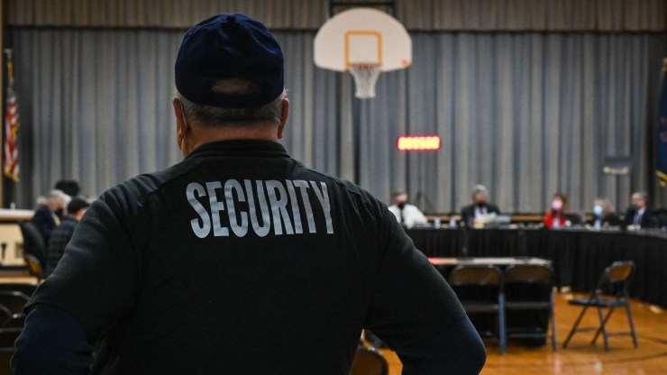 The back of a security guard, who wears a black baseball cap and long-sleeve shirt that reads "SECURITY" is seen in front a long table in a gymnasium, where a school board meeting is taking place.